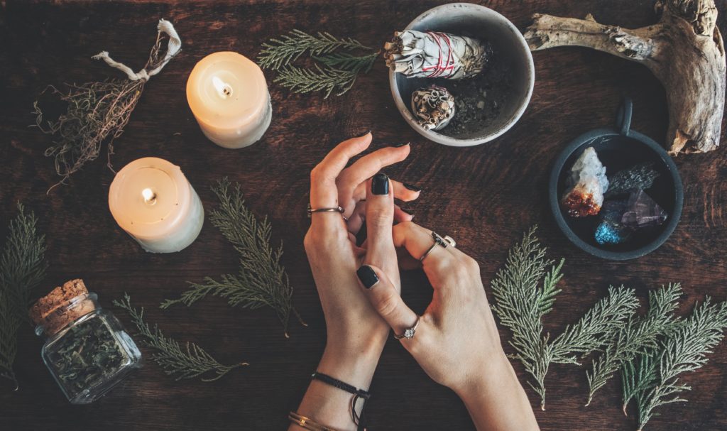 Witches hands on a table ready for spell work. Wiccan witch altar filled with sage evergreen branches herbs crystals and burning white candles. Wearing vintage jewelry on her hands