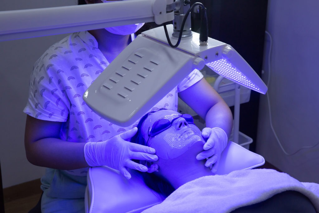 Young woman having blue LED light facial therapy treatment in beauty salon. Beautician wearing face mask maintaining safety procedures during appointment. Beauty, new normal and wellness