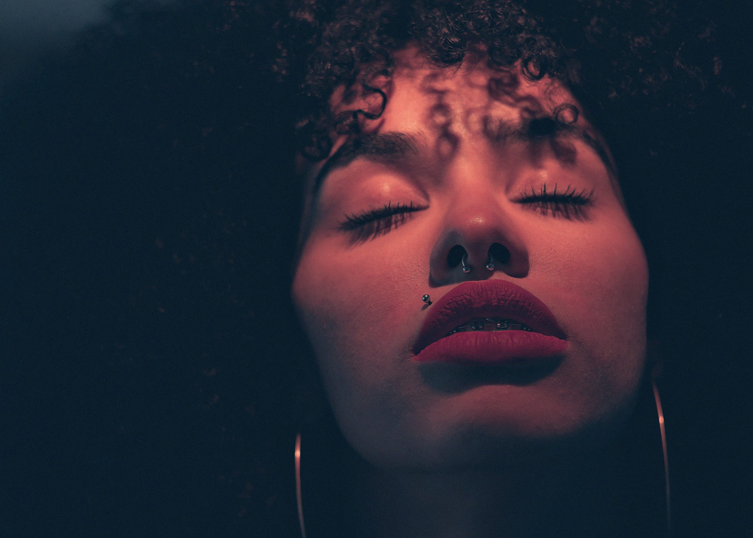 a woman with a nose ring and a Madonna lip piercing with her head raised and eyes closed under a red light