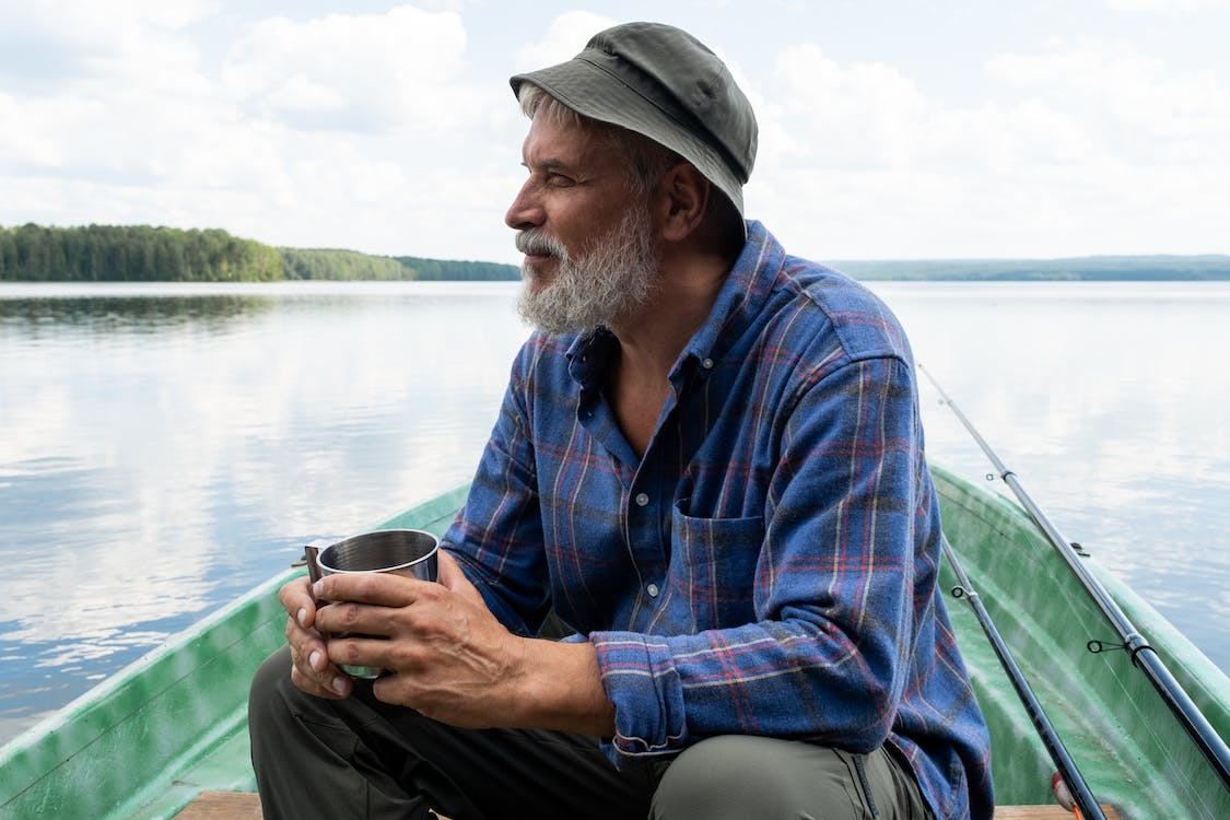 Bearded Senior Man in Hat Sitting in Boat and Holding Mug  